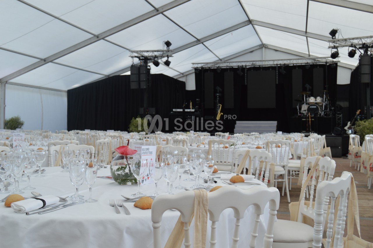 Salle de réception sous chapiteau avec tables dressées, nappes blanches, verres, couverts et scène équipée en arrière-plan.