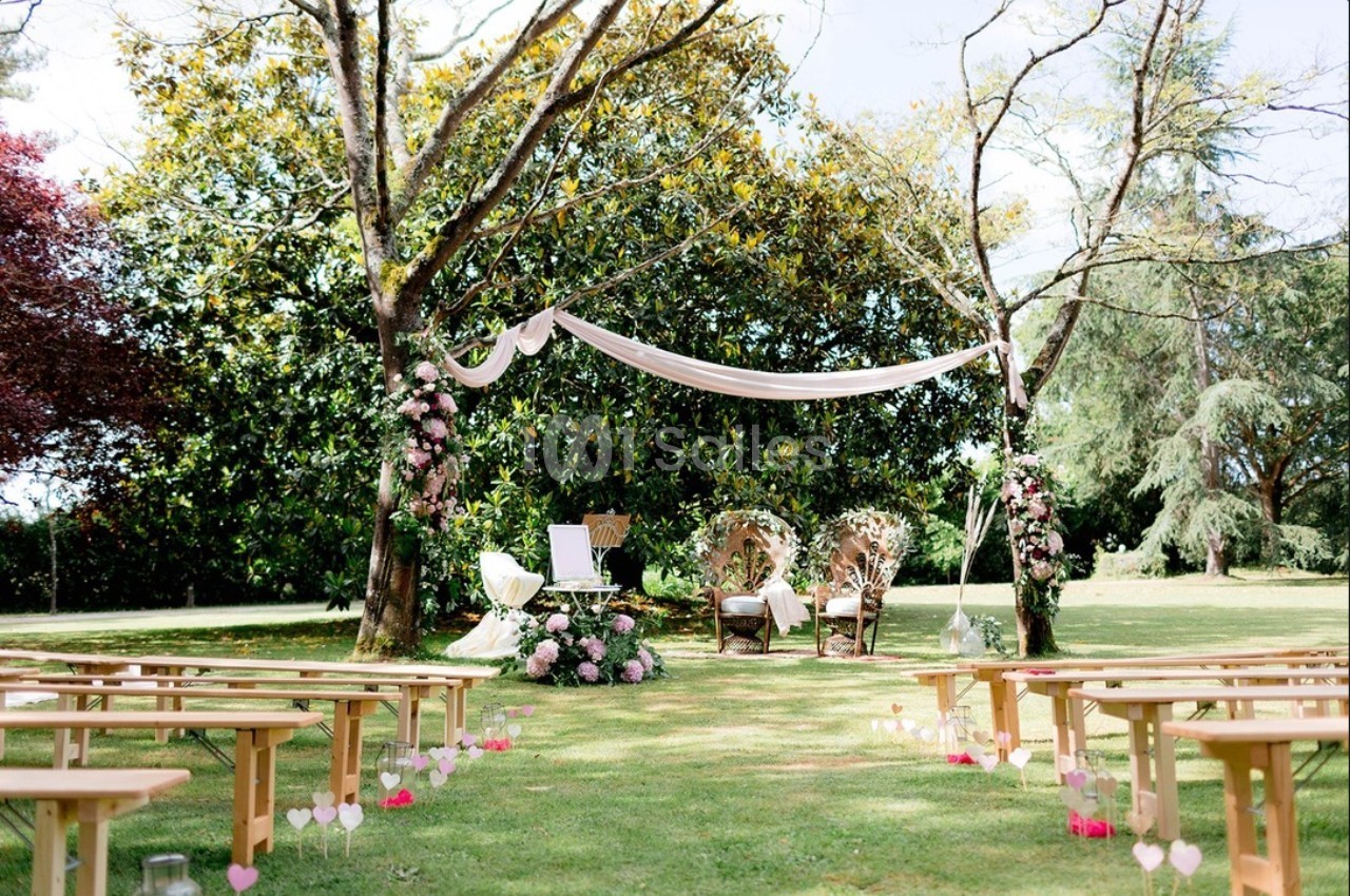 Cérémonie en plein air avec arche décorée de fleurs, chaises en rotin et bancs en bois disposés sur une pelouse.
