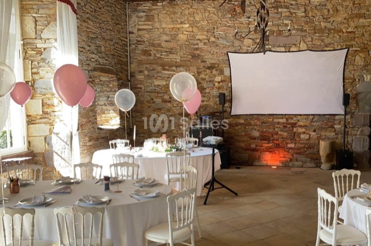 Salle en pierre décorée pour un événement, avec tables dressées, ballons roses et blancs, et un écran de projection.