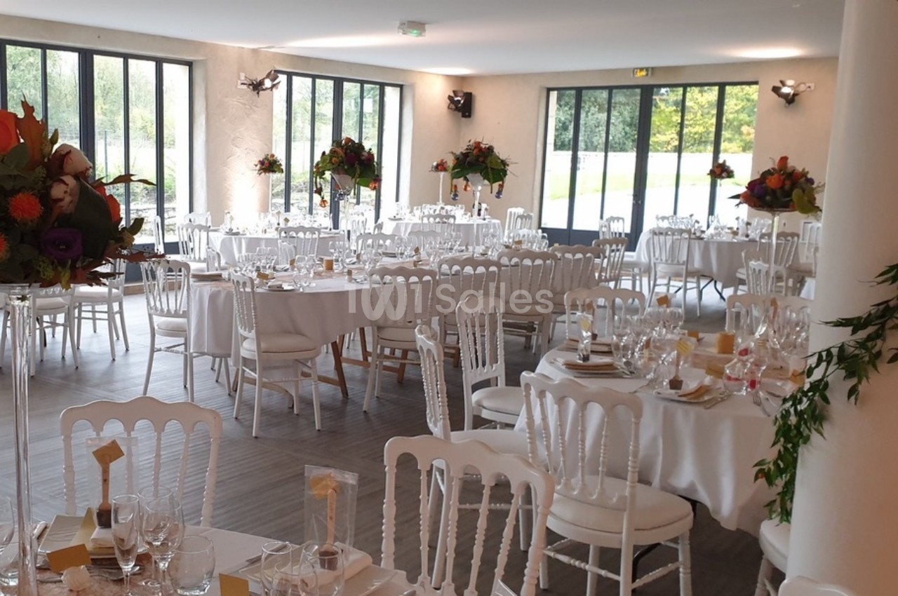 Salle de réception lumineuse avec tables rondes dressées, chaises blanches et décorations florales.