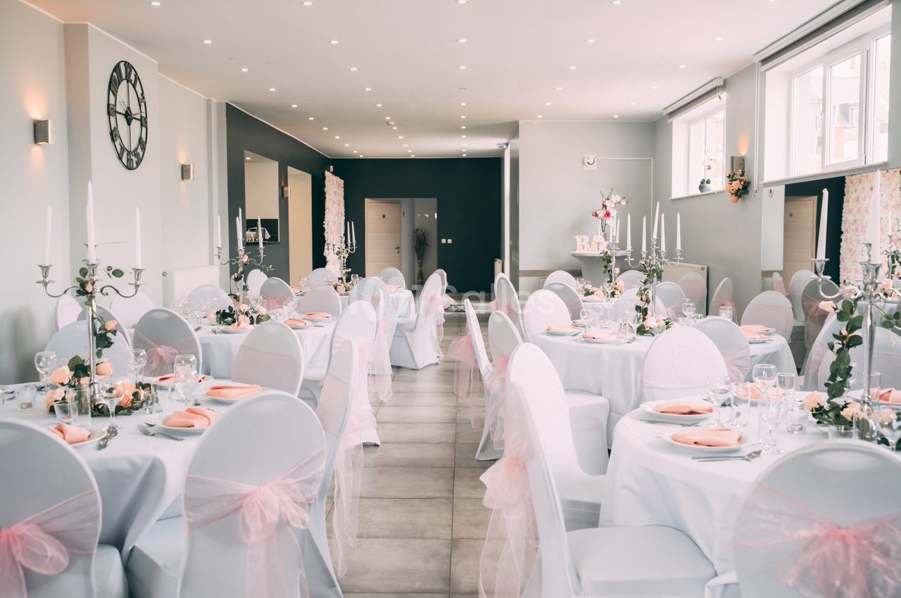 Salle de réception décorée avec des tables rondes, nappes blanches, chaises ornées de nœuds roses et chandeliers.