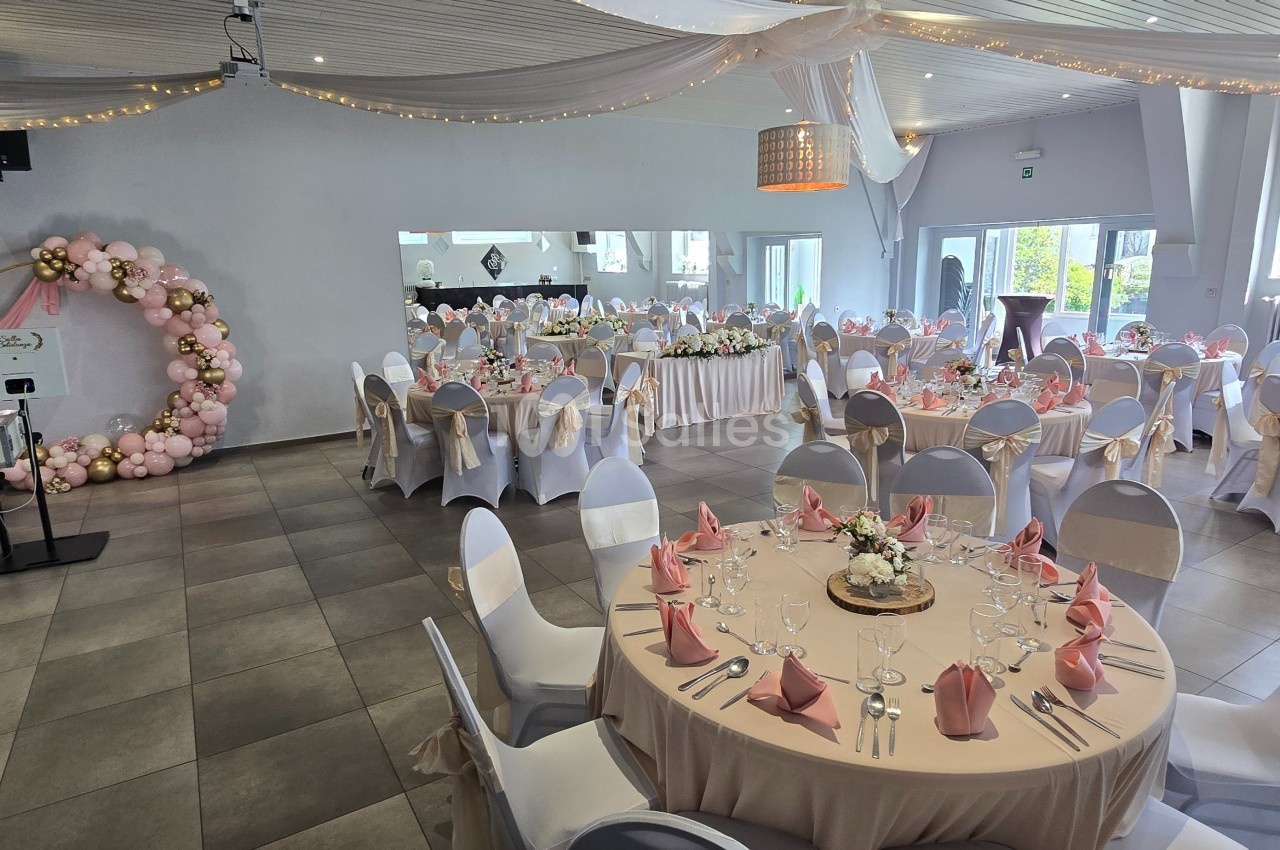 Salle de réception décorée pour un événement, avec tables rondes dressées, nappes blanches et accents roses.