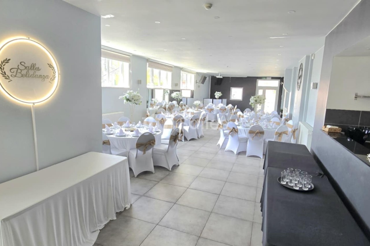 Salle de réception décorée avec des tables rondes, nappes blanches et chaises ornées de nœuds beiges.