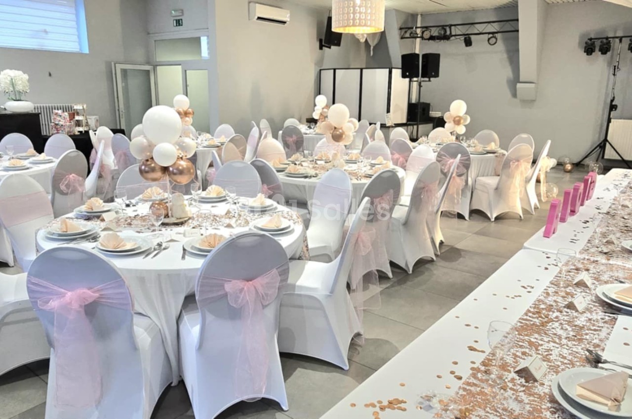 Salle de réception décorée pour un événement, avec tables rondes dressées, nappes blanches et décorations roses et dorées.