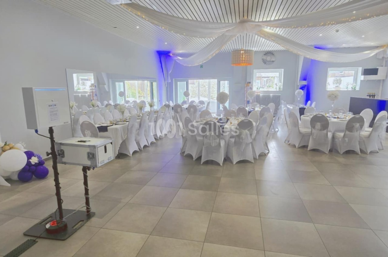 Salle de réception décorée avec des tables rondes dressées, des chaises blanches et des ballons en arrière-plan.