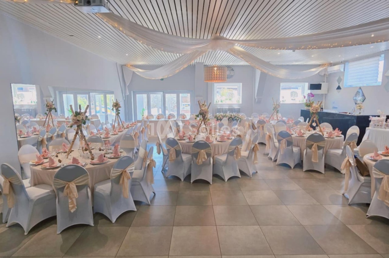 Salle de réception décorée avec des tables rondes, nappes blanches, chaises ornées de nœuds et éclairage chaleureux.