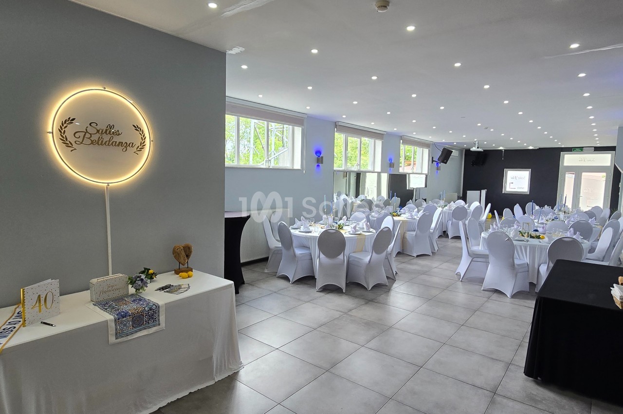 Salle de réception décorée avec des tables rondes dressées, chaises blanches et éclairage lumineux.