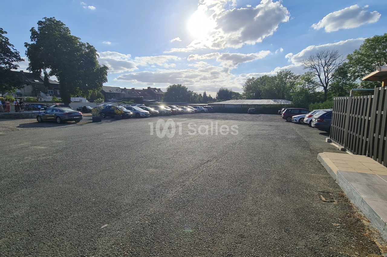 Parking extérieur en gravier avec plusieurs voitures stationnées, entouré d'arbres et de bâtiments sous un ciel ensoleillé.