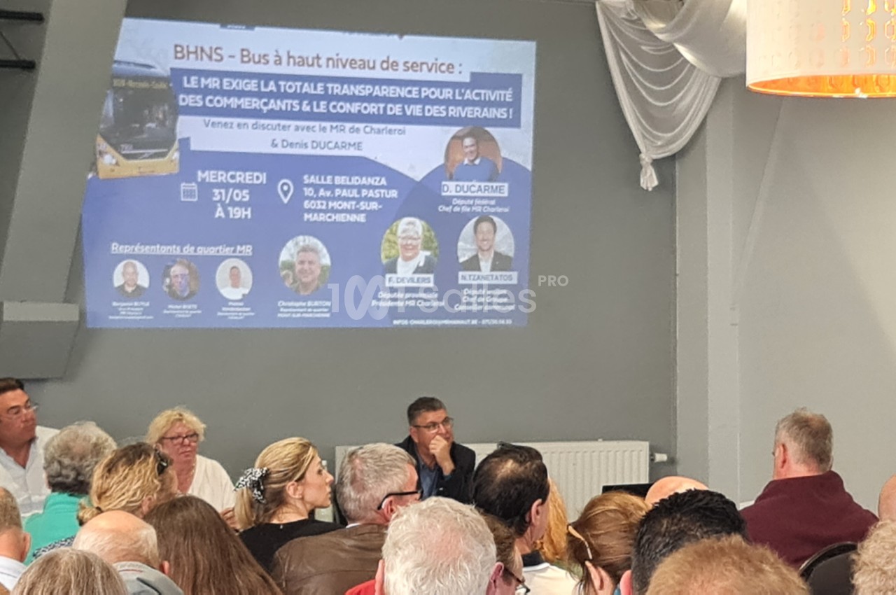 Un groupe de personnes assiste à une réunion publique dans une salle, avec une présentation projetée au mur.