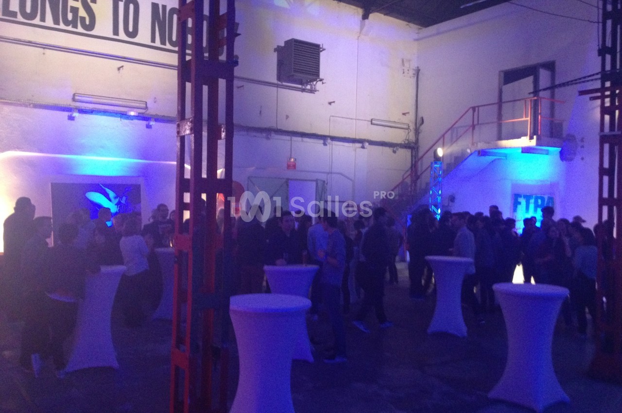 Des personnes rassemblées dans un espace industriel éclairé par des lumières colorées, avec des tables hautes blanches.