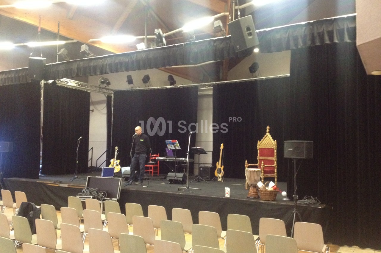 Scène équipée de micros, instruments et fauteuil, dans une salle avec des rangées de chaises vides.