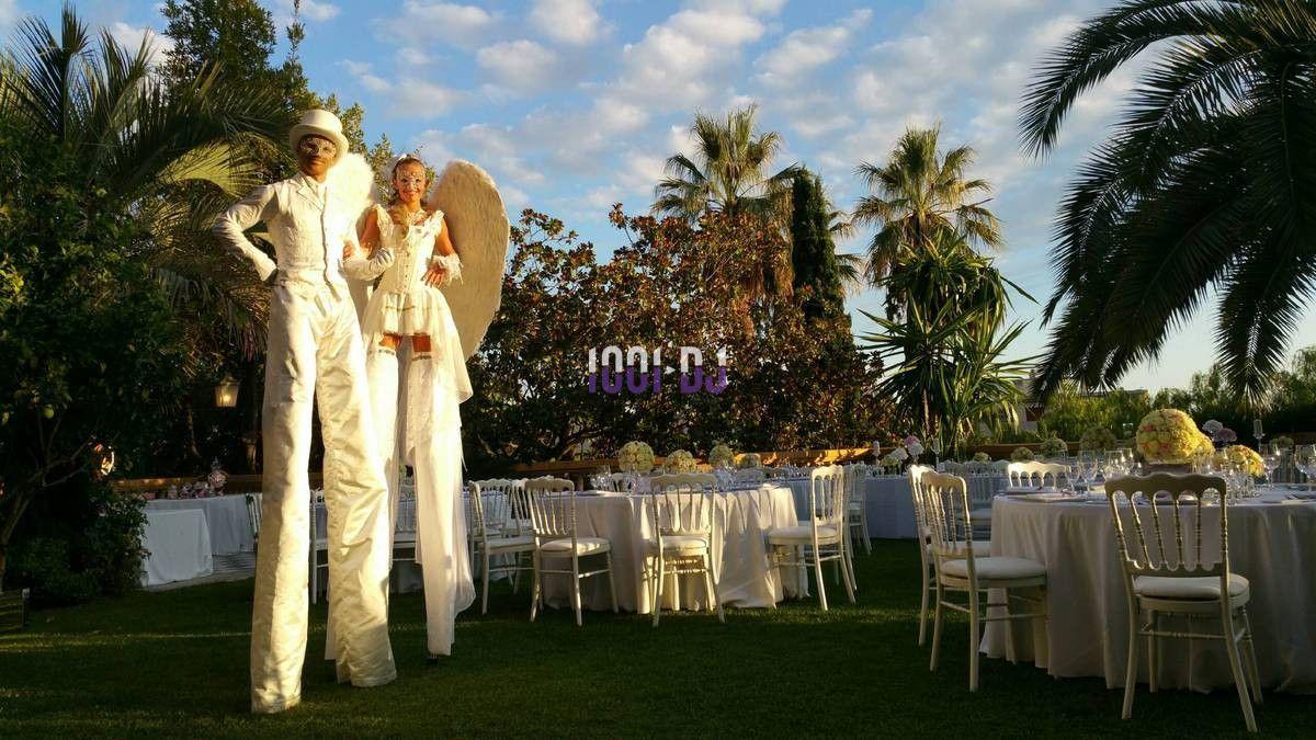 Deux artistes sur échasses en costumes blancs dans un jardin, entourés de tables dressées pour un événement en plein air.