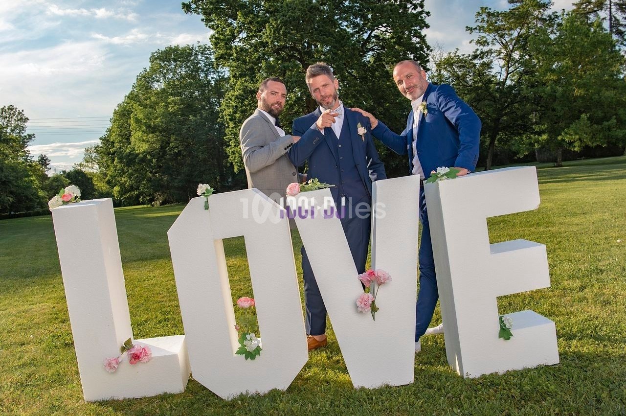 Trois hommes en costume posent devant de grandes lettres blanches formant le mot ’LOVE’ dans un parc verdoyant.