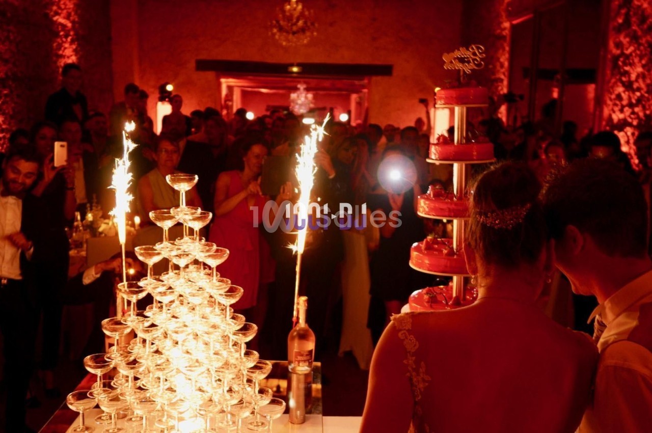 Un couple de mariés observe une fontaine de champagne et un gâteau illuminés par des cierges dans une salle festive.