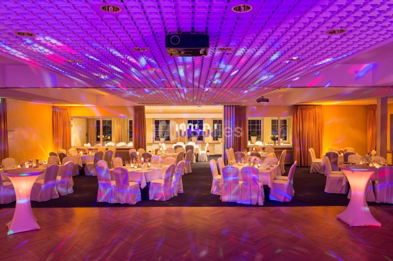 Salle de réception éclairée par des lumières colorées, avec tables dressées et chaises recouvertes de housses blanches.