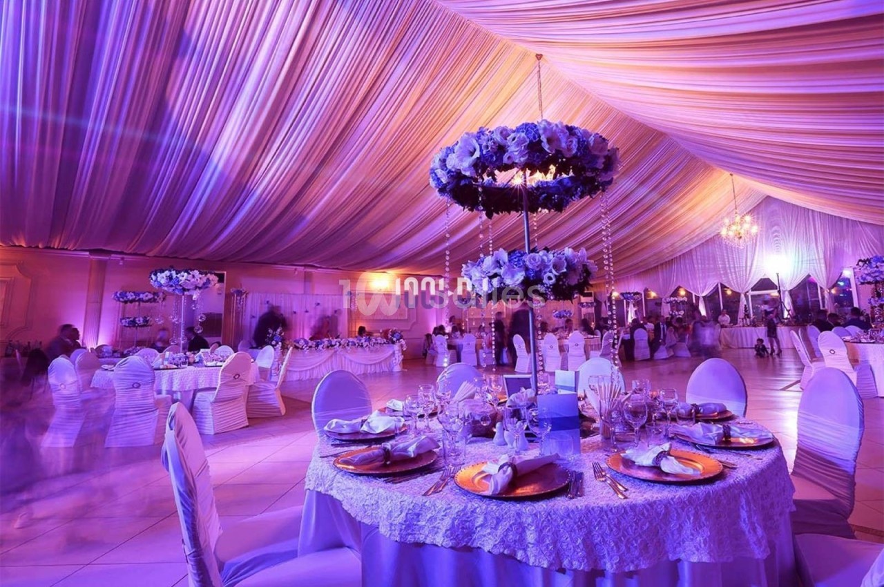 Salle de réception décorée avec des tables rondes élégantes, des fleurs suspendues et un éclairage violet tamisé.
