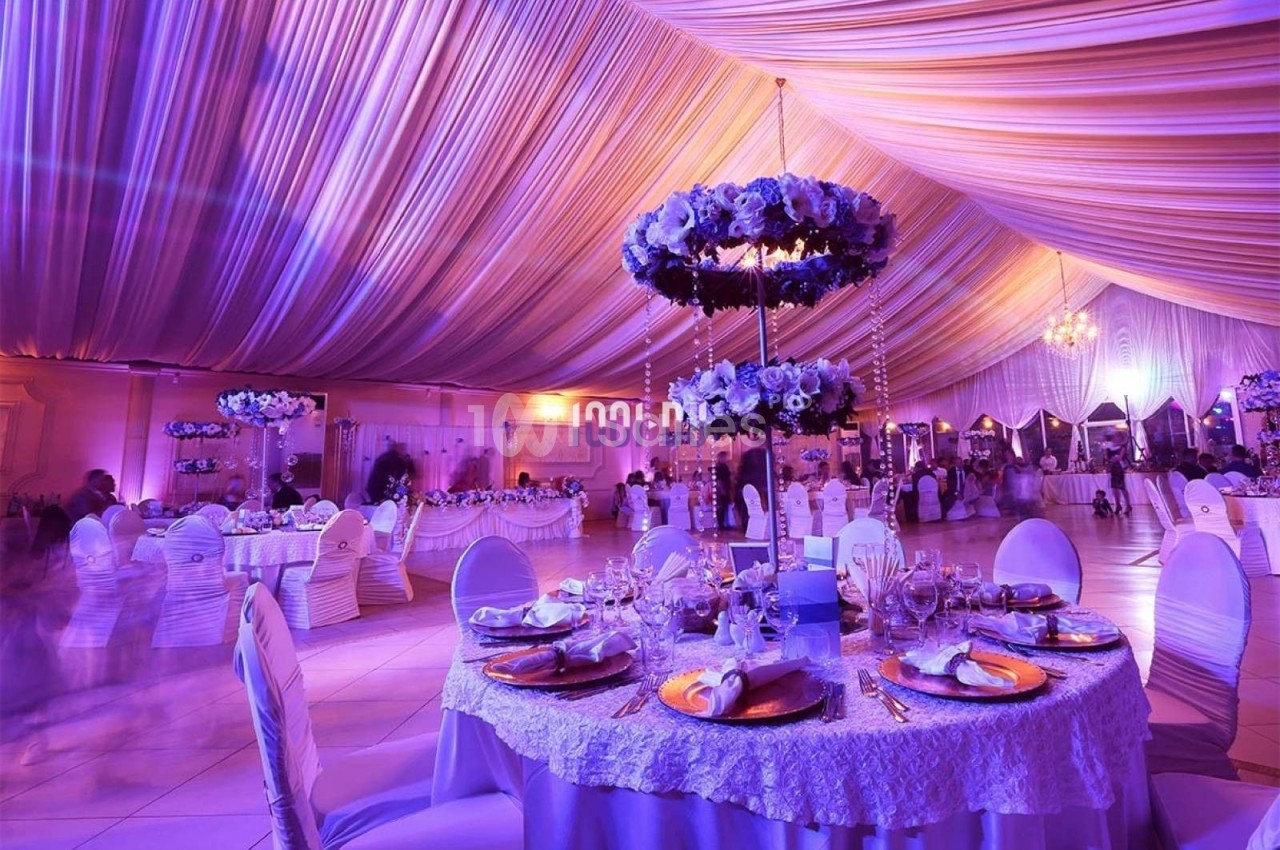 Salle de réception décorée avec des tables rondes élégantes, des fleurs suspendues et un éclairage violet tamisé.