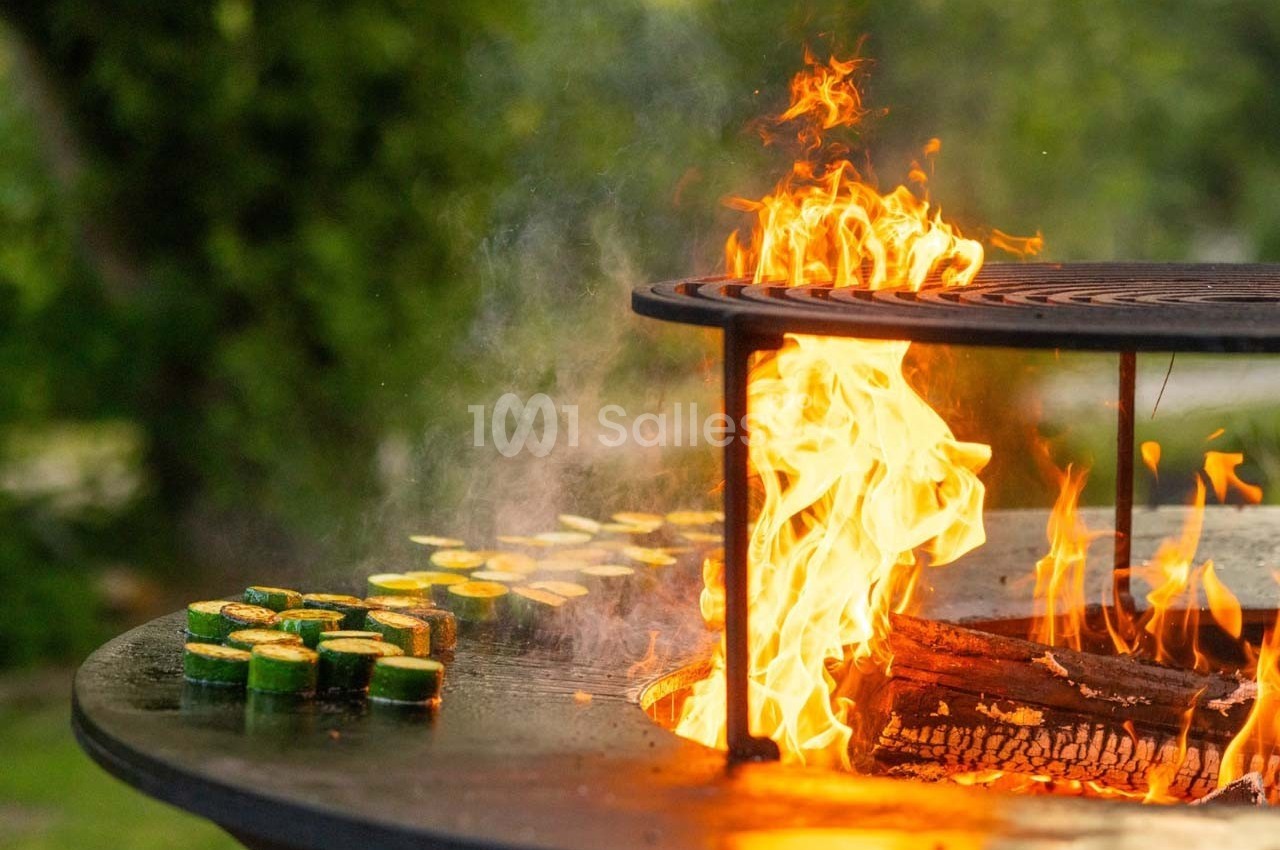 Légumes grillant sur un barbecue circulaire avec des flammes vives en extérieur verdoyant.