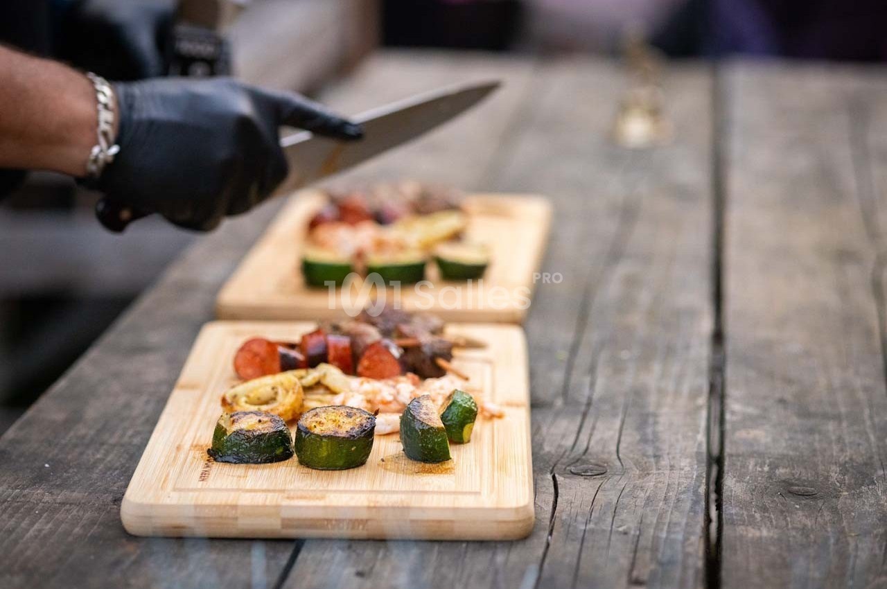 Planche en bois garnie de légumes grillés, saucisses et viande, avec une main coupant des aliments au couteau.