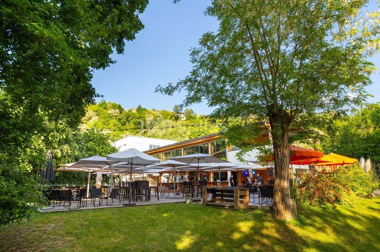 Terrasse d'un restaurant entourée de verdure, avec des tables, des parasols et un bâtiment moderne en arrière-plan.