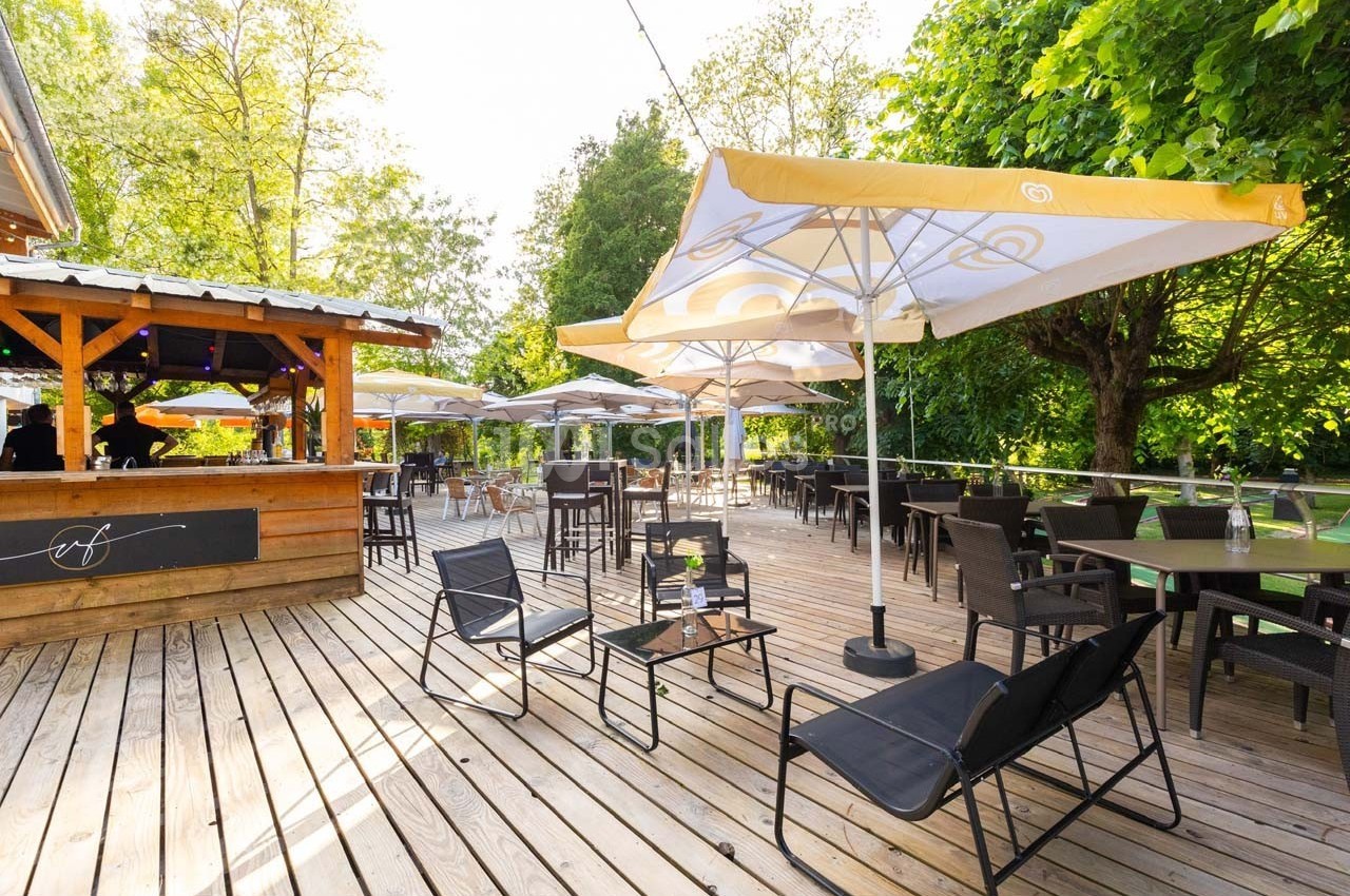 Terrasse en bois avec tables, chaises et parasols jaunes, entourée de verdure sous un ciel dégagé.