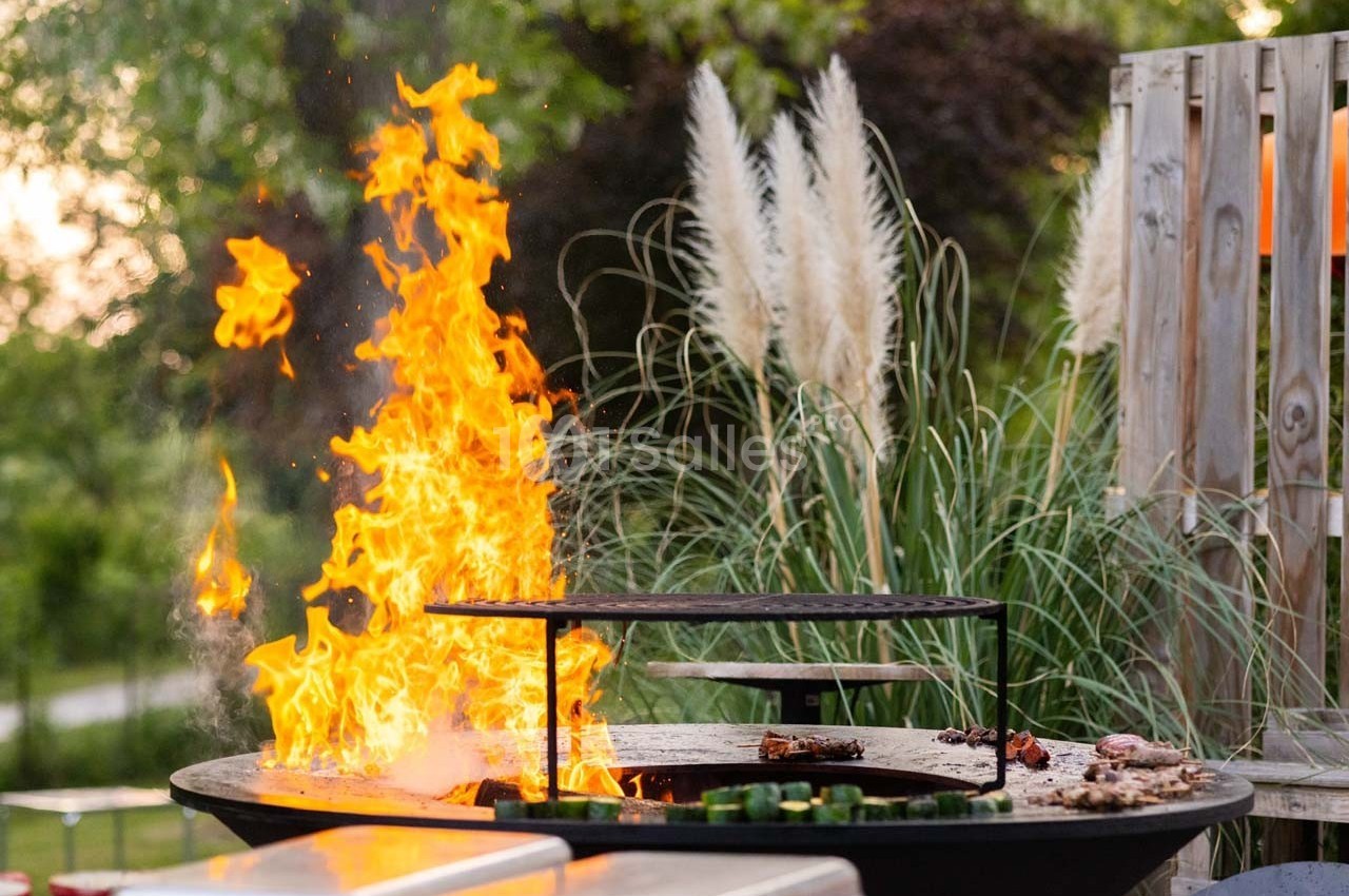 Barbecue circulaire en plein air avec des flammes vives, entouré de végétation et de mobilier de jardin.