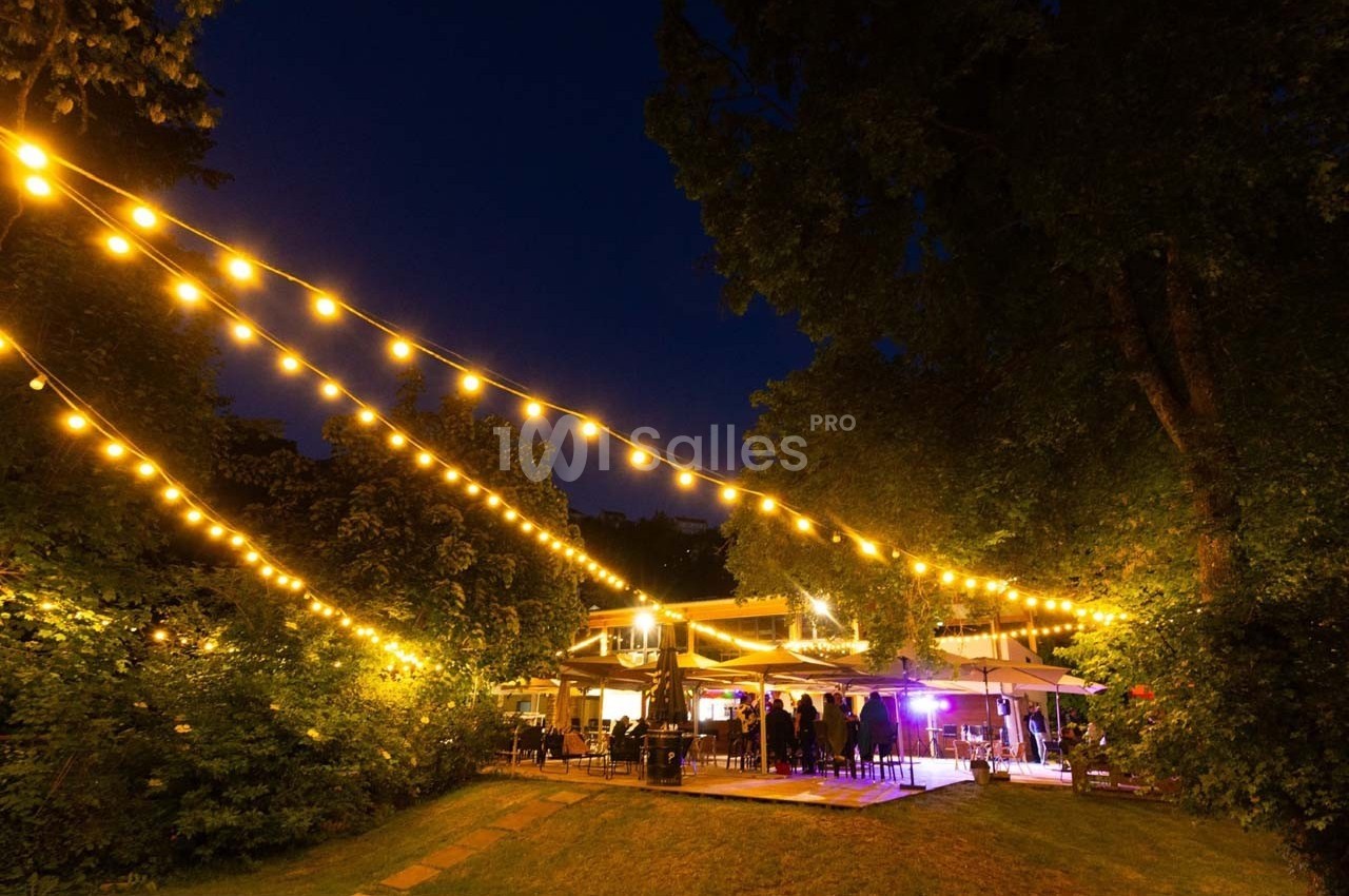 Terrasse extérieure éclairée par des guirlandes lumineuses, entourée d'arbres, avec des personnes rassemblées en soirée.