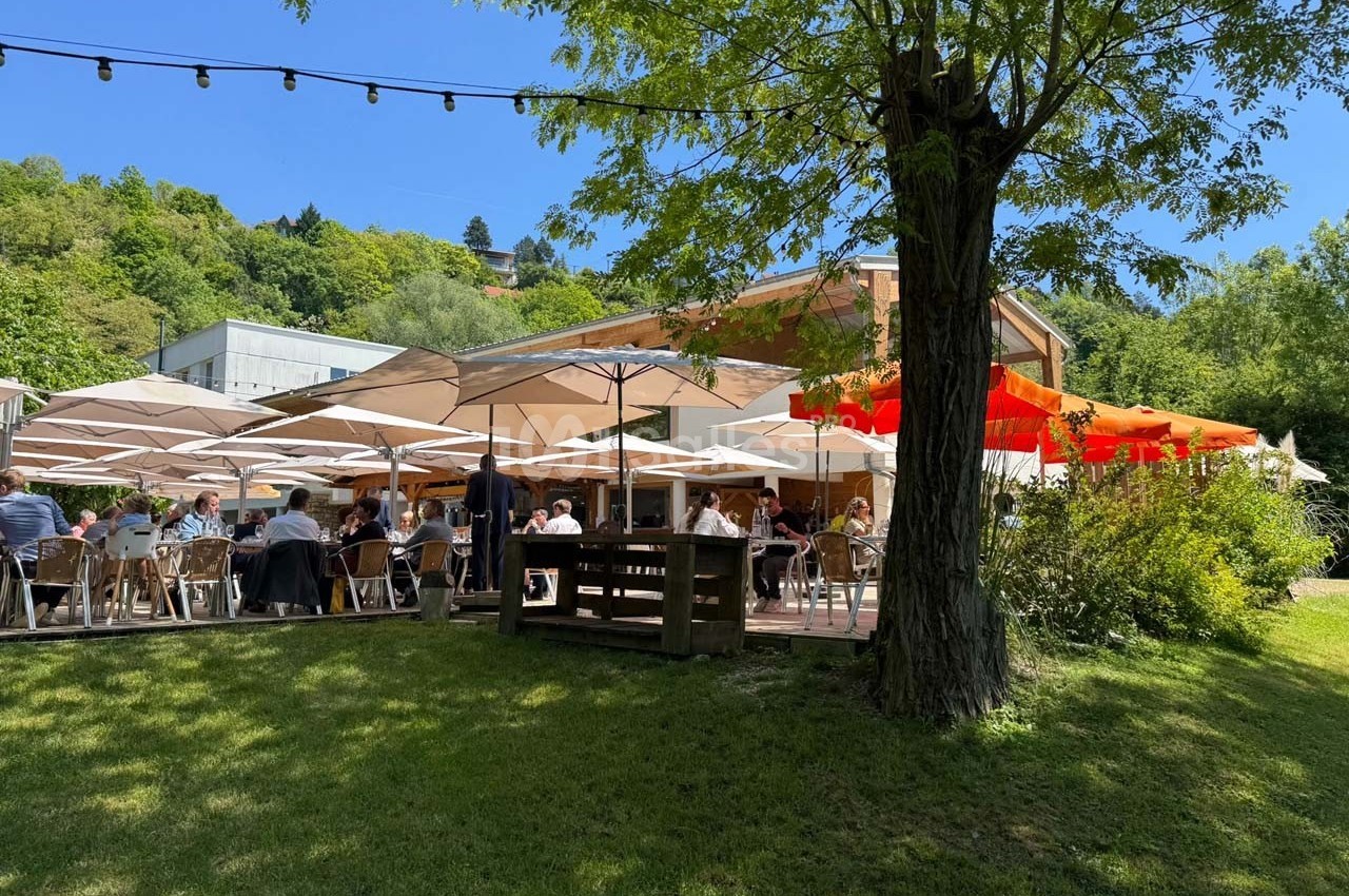 Terrasse extérieure d'un restaurant avec parasols, tables occupées et arbres environnants dans un cadre verdoyant.
