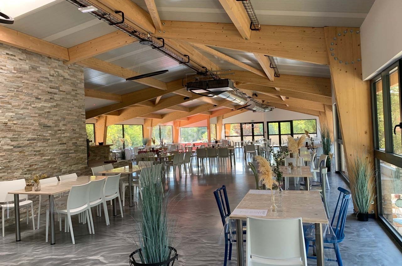 Salle lumineuse avec charpente en bois, tables et chaises modernes, grandes baies vitrées donnant sur un espace arboré.