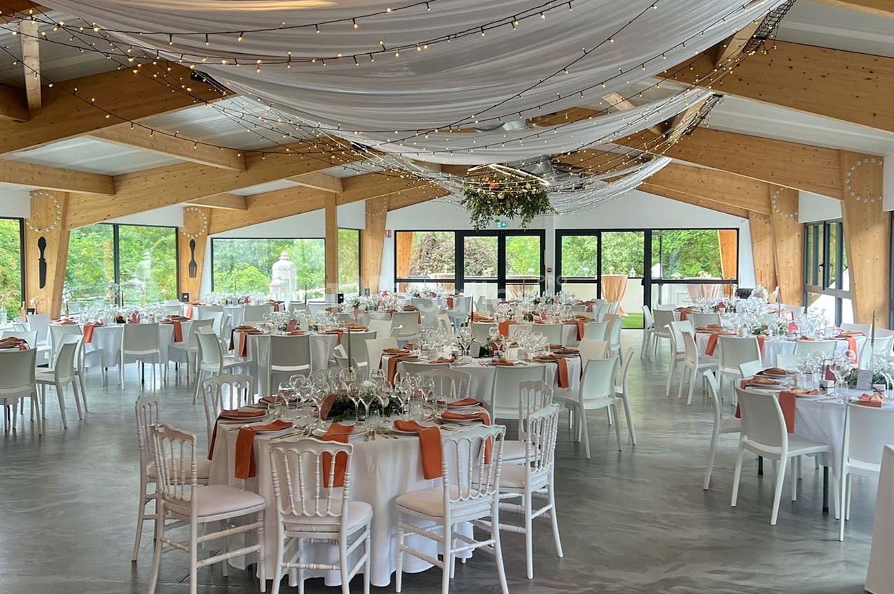 Salle de réception décorée avec des tables rondes dressées, nappes blanches, chaises blanches et guirlandes lumineuses…