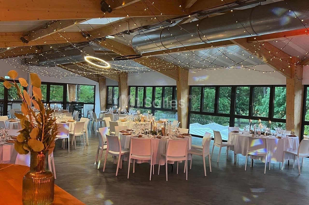 Salle de réception lumineuse avec tables dressées, grandes baies vitrées et guirlandes lumineuses suspendues.