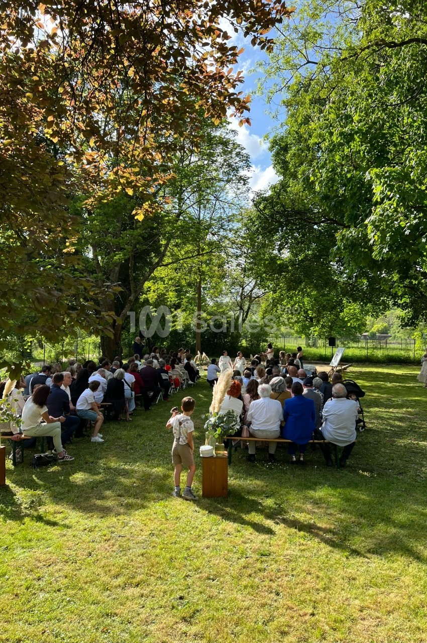 Cérémonie en plein air avec des invités assis sur des bancs dans un jardin verdoyant sous un ciel ensoleillé.