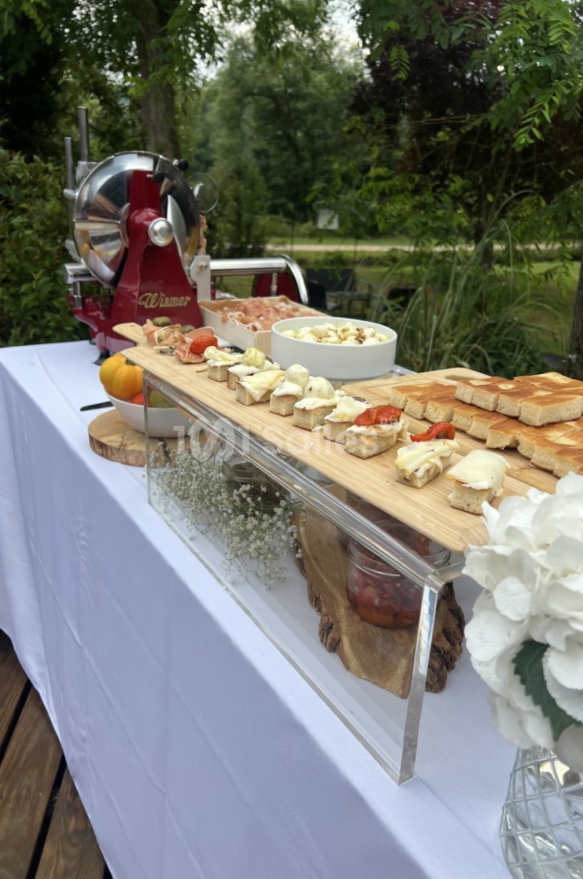 Plateau de toasts et amuse-bouches disposés sur une table blanche en extérieur, avec une trancheuse et un décor végétal.