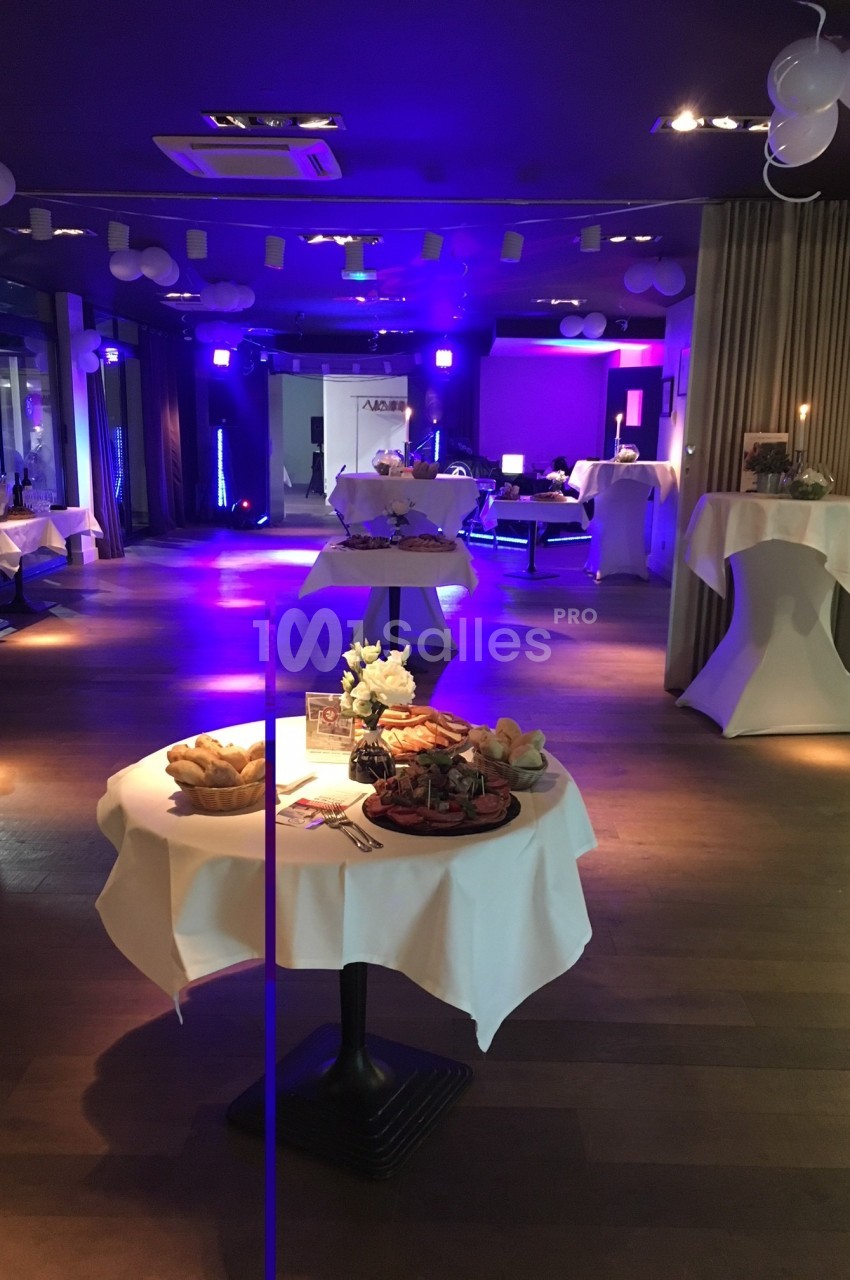 Salle décorée pour un événement avec tables rondes, nappes blanches, bougies et éclairage tamisé.
