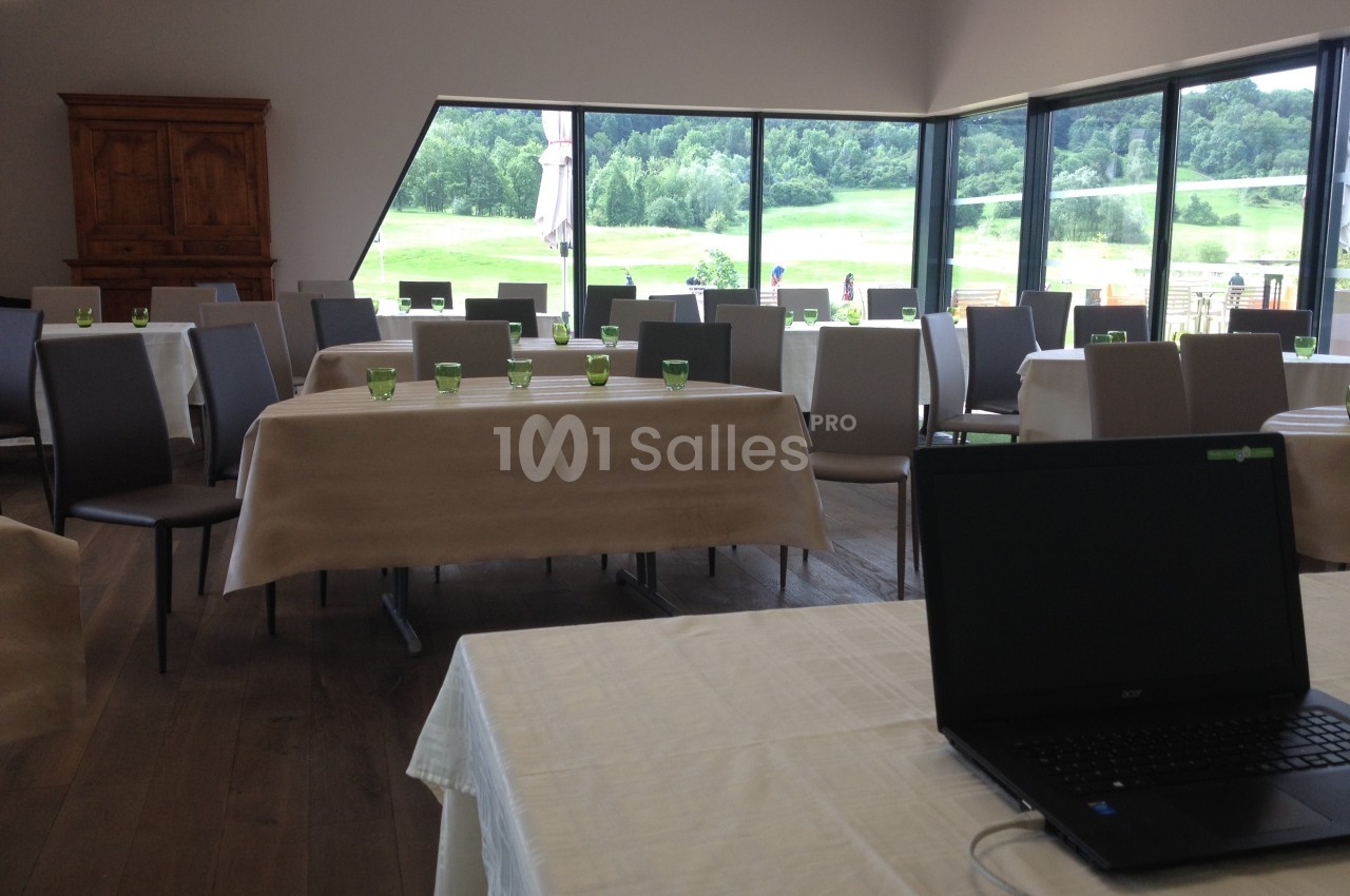 Salle lumineuse avec tables dressées, chaises modernes, grandes baies vitrées donnant sur un paysage verdoyant.