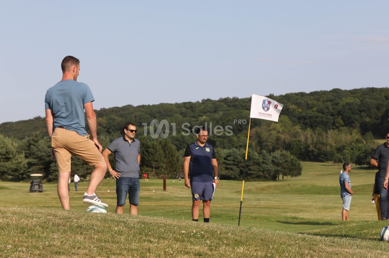 Un homme joue au football sur un terrain herbeux, observé par trois personnes près d'un drapeau.