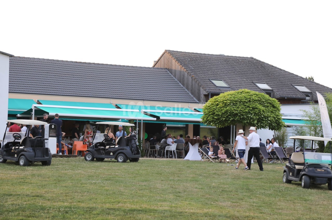 Terrasse d'un bâtiment avec des tables occupées, des personnes debout et des voiturettes de golf stationnées sur la pelouse.