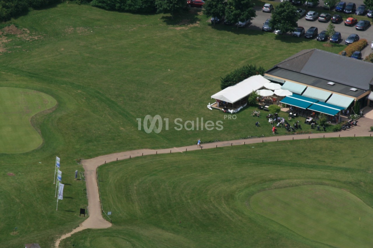 Vue aérienne d'un terrain de golf avec un bâtiment, une terrasse couverte et des voitures garées à proximité.