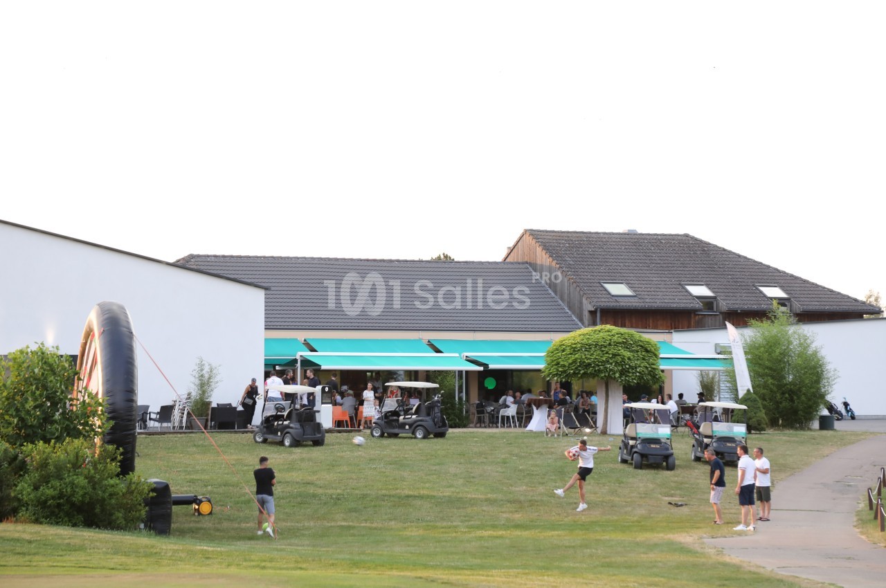 Des personnes rassemblées devant un bâtiment moderne avec des terrasses, des parasols et des voiturettes de golf.
