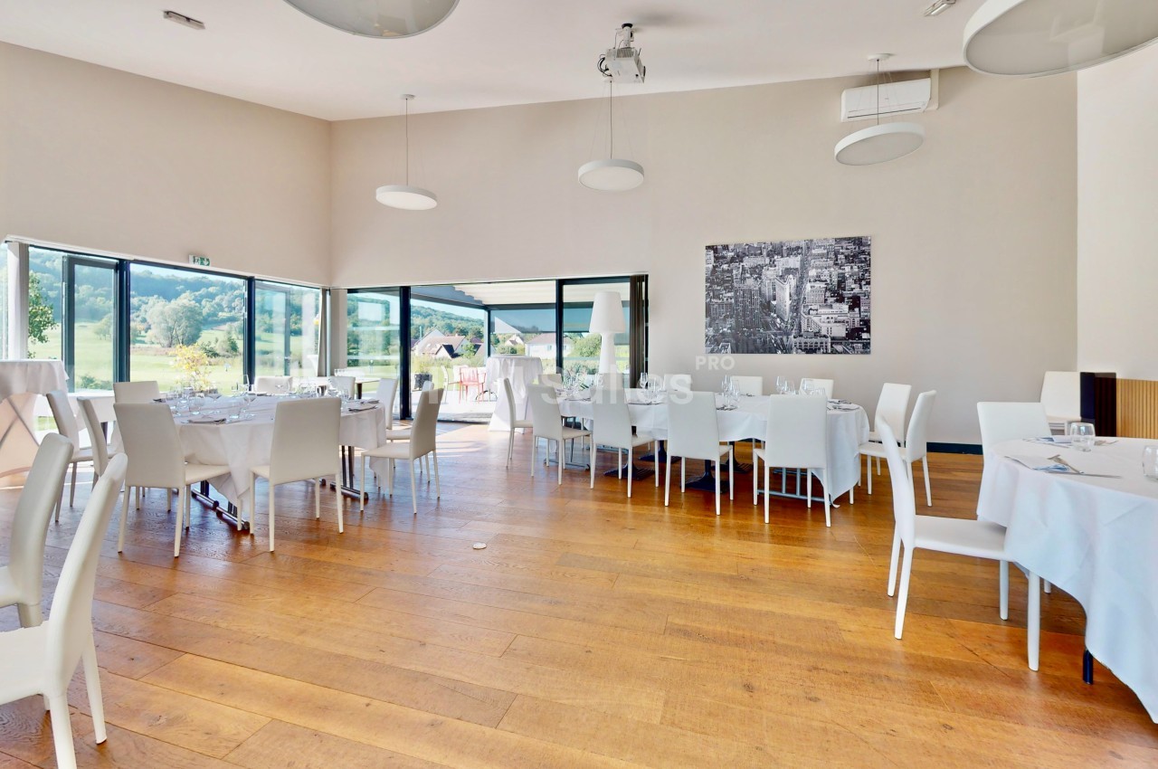 Salle lumineuse avec parquet en bois, tables dressées avec nappes blanches et grandes baies vitrées donnant sur l'extérieur.