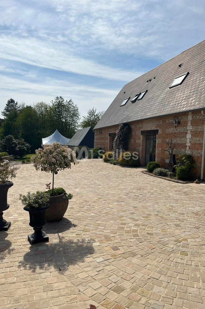 Cour pavée devant une maison en briques avec toit en ardoise, entourée de végétation et de pots de fleurs.