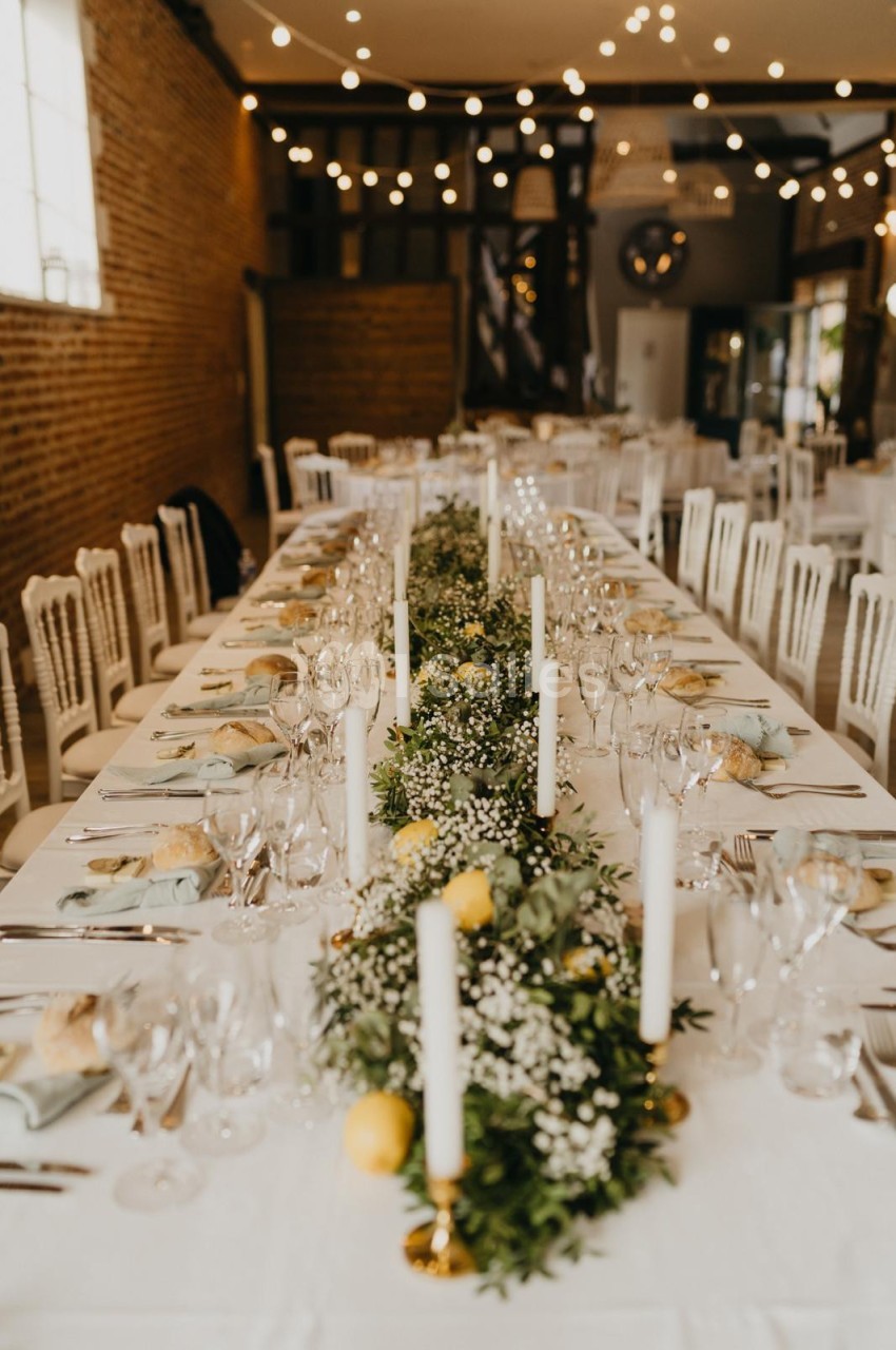 Table de réception décorée avec des bougies, des fleurs blanches et des feuillages, dans une salle éclairée par des…