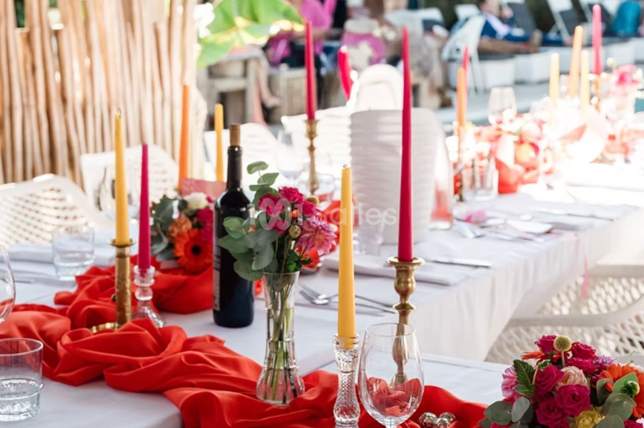 Table décorée avec des nappes rouges, chandeliers, bougies colorées, fleurs et une bouteille de vin dans un cadre extérieur.