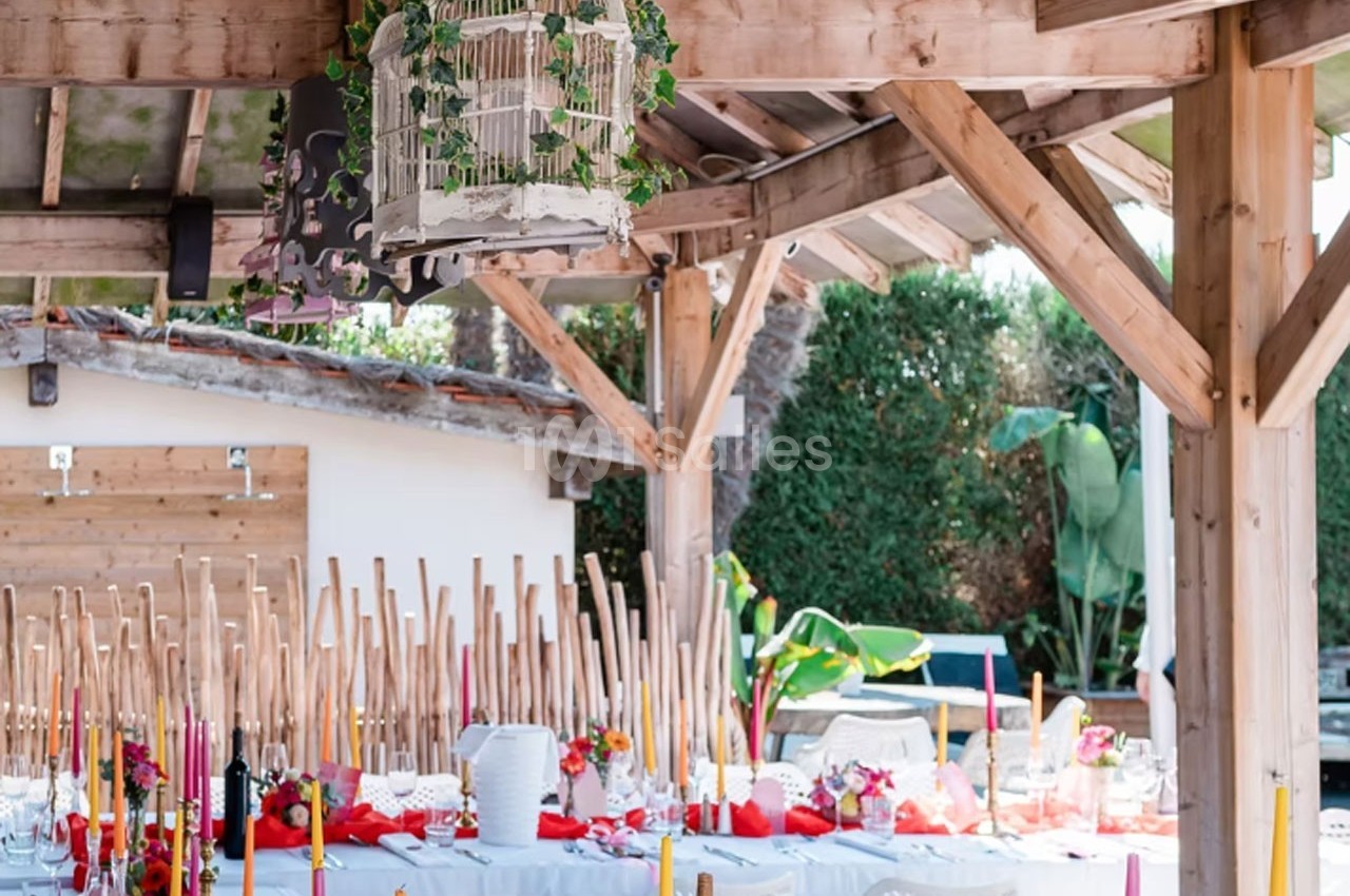 Salle de réception extérieure décorée avec des tables dressées, des chandeliers colorés et une cage suspendue ornée de…