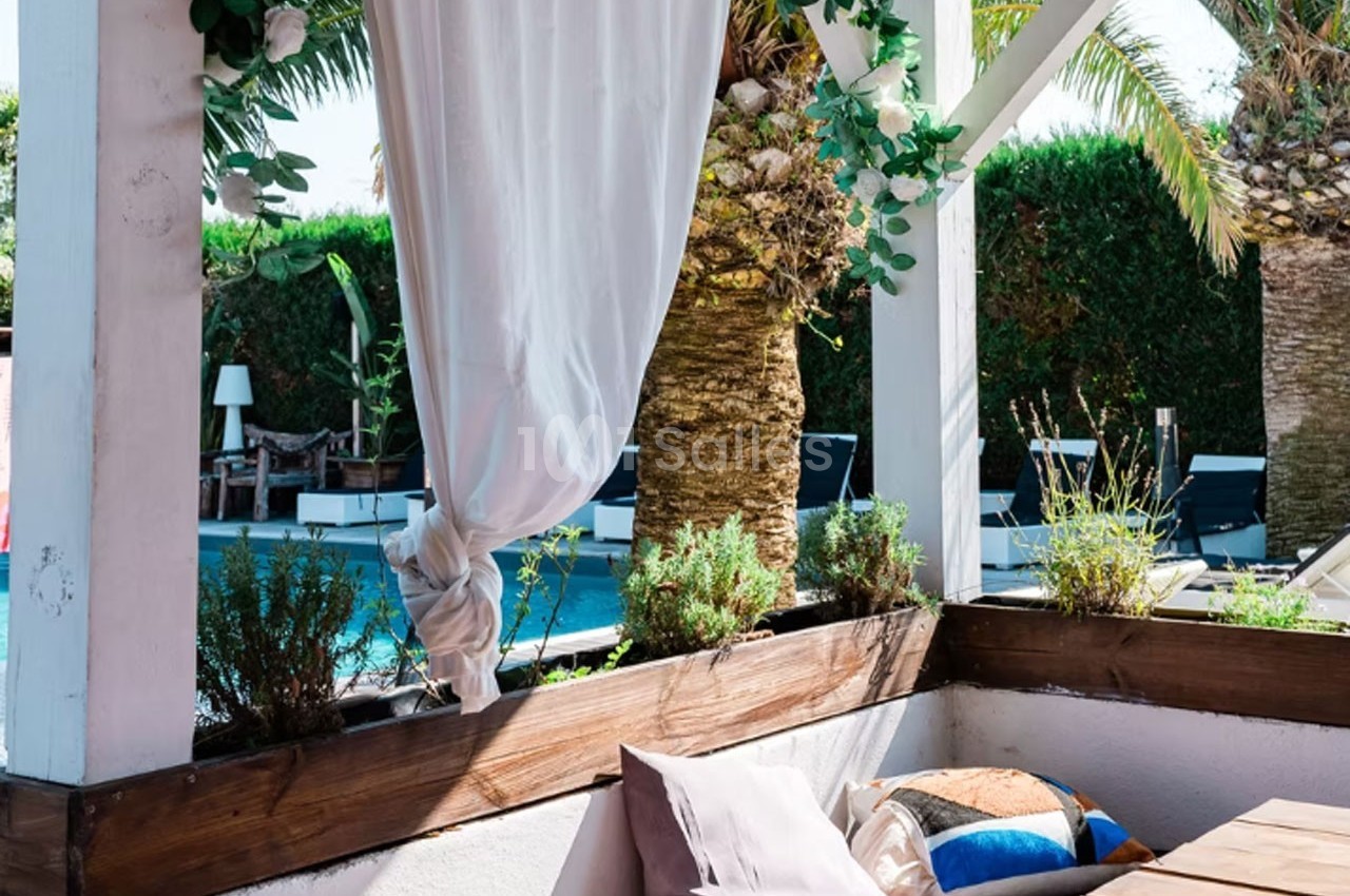 Terrasse ombragée avec rideaux blancs, coussins colorés, plantes en jardinières et vue sur une piscine entourée de palmiers.