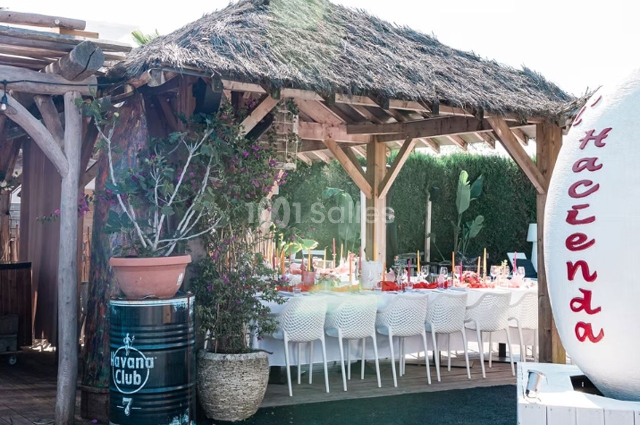 Table dressée sous une pergola en bois avec toit en chaume, entourée de plantes et d'éléments décoratifs extérieurs.