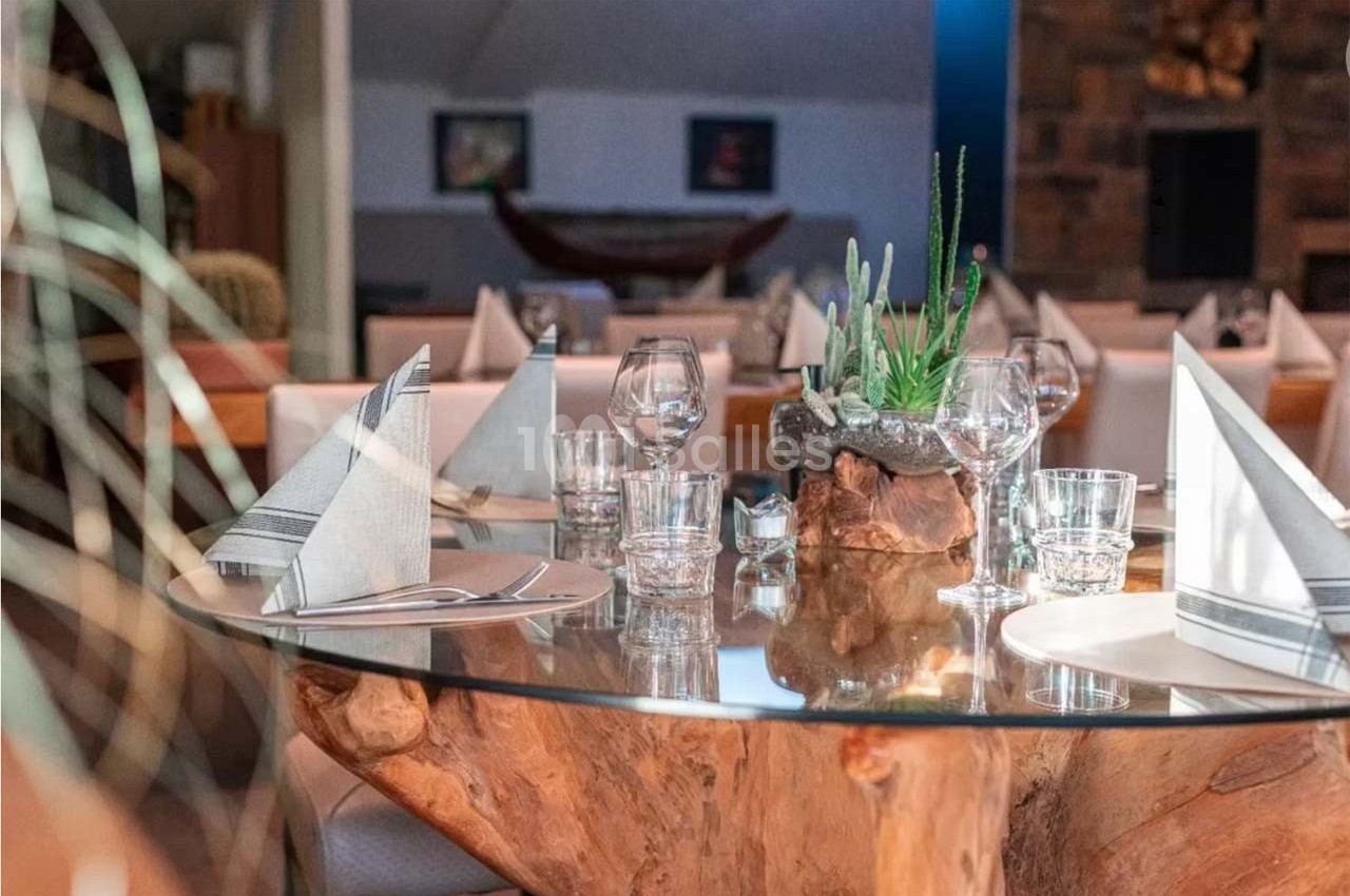 Table en verre avec pied en bois sculpté, dressée avec serviettes pliées, verres et assiettes dans un restaurant élégant.