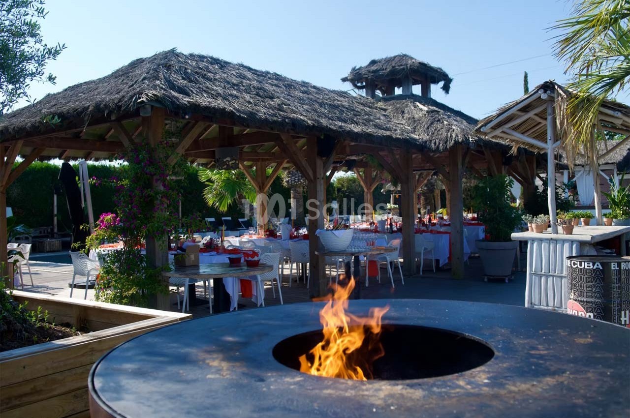Terrasse extérieure avec tables dressées sous des toits en chaume, et un brasero allumé au premier plan.