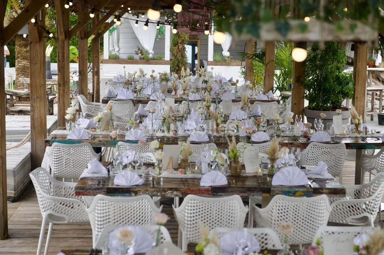Tables décorées pour un événement, disposées sous une pergola en bois avec des chaises blanches et des guirlandes lumineuses.