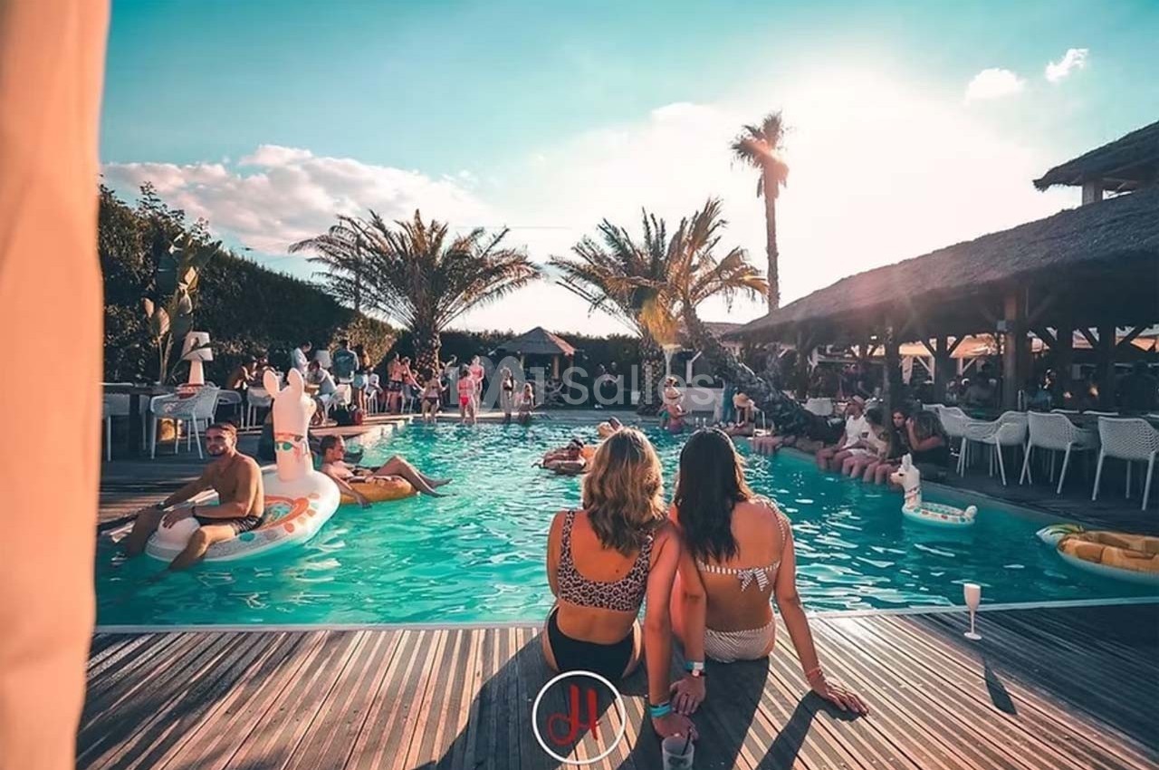 Des personnes profitent d'une piscine entourée de palmiers et de terrasses en bois sous un ciel ensoleillé.